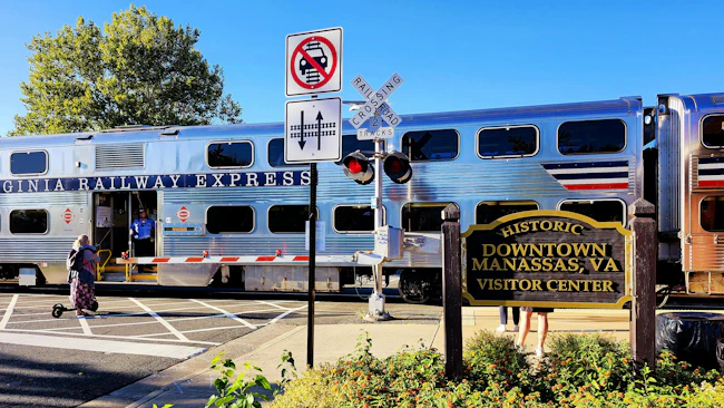 A silver Virginia Railway Express bilevel commuter rail car with red, white and blue striping sits at a grade crossing with a conductor visible in the open doorway, with a 'Historic Downtown Manassas, VA Visitor Center' sign and railroad crossing signals in the foreground.