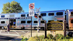 A silver Virginia Railway Express bilevel commuter rail car with red, white and blue striping sits at a grade crossing with a conductor visible in the open doorway, with a 'Historic Downtown Manassas, VA Visitor Center' sign and railroad crossing signals in the foreground.