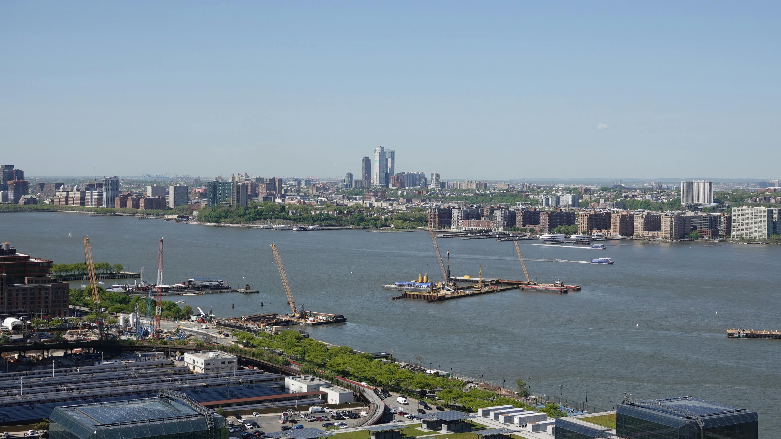 Construction platforms, cranes and barges sit on the Hudson River off Manhattan's West Side as work continues on the Gateway Program's Hudson Tunnel Project, with the Javits Center's green roof in the foreground and the Jersey City and Hoboken skylines across the water.