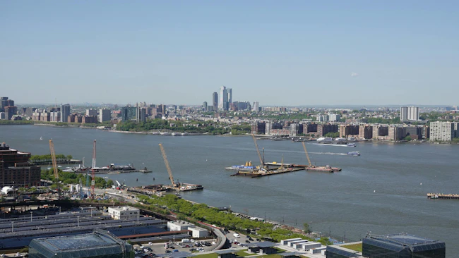 Construction platforms, cranes and barges sit on the Hudson River off Manhattan's West Side as work continues on the Gateway Program's Hudson Tunnel Project, with the Javits Center's green roof in the foreground and the Jersey City and Hoboken skylines across the water.