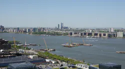 Construction platforms, cranes and barges sit on the Hudson River off Manhattan's West Side as work continues on the Gateway Program's Hudson Tunnel Project, with the Javits Center's green roof in the foreground and the Jersey City and Hoboken skylines across the water.