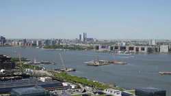 Construction platforms, cranes and barges sit on the Hudson River off Manhattan's West Side as work continues on the Gateway Program's Hudson Tunnel Project, with the Javits Center's green roof in the foreground and the Jersey City and Hoboken skylines across the water.