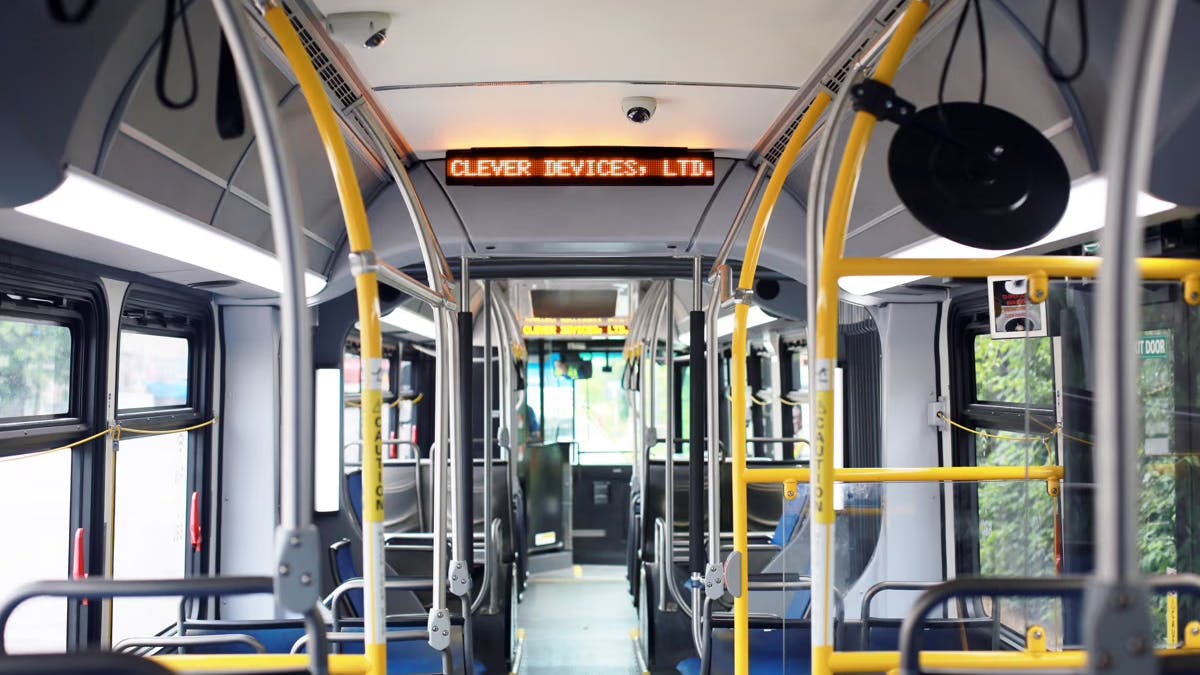 The interior of a transit bus with yellow stanchions and blue seating, showing an orange LED destination sign reading 'Clever Devices, Ltd.' with a matching display visible through the bus at the front destination sign as well.