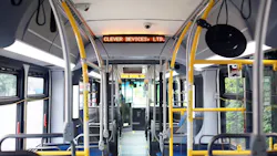 The interior of a transit bus with yellow stanchions and blue seating, showing an orange LED destination sign reading 'Clever Devices, Ltd.' with a matching display visible through the bus at the front destination sign as well.