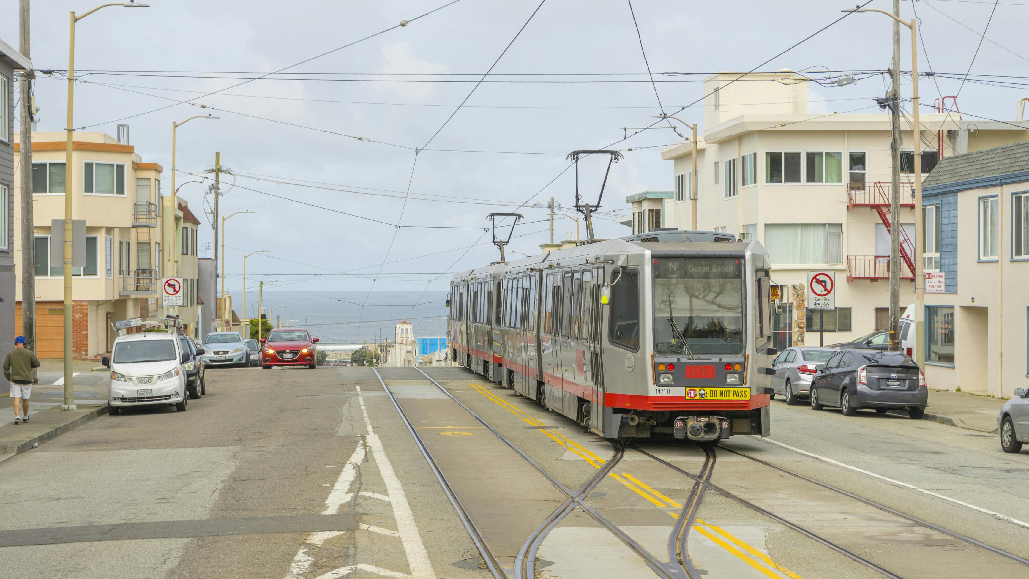A silver and red San Francisco Municipal Transportation Agency Muni light rail vehicle operating the N Judah line to Ocean Beach runs along street-level tracks in San Francisco's Outer Sunset neighborhood, with the Pacific Ocean visible at the end of the street under an overcast sky.