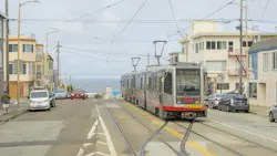 A silver and red San Francisco Municipal Transportation Agency Muni light rail vehicle operating the N Judah line to Ocean Beach runs along street-level tracks in San Francisco's Outer Sunset neighborhood, with the Pacific Ocean visible at the end of the street under an overcast sky.