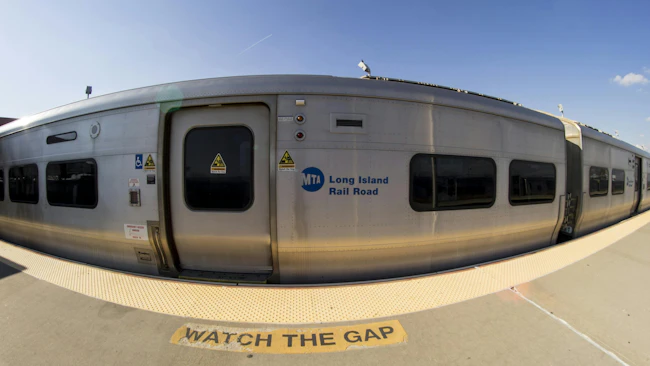 A fisheye lens view of a silver MTA Long Island Rail Road train stopped at a station platform, with 'Watch the Gap' warning painted on the concrete in front of the open doorway and the agency's blue circular logo visible on the car side.