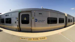 A fisheye lens view of a silver MTA Long Island Rail Road train stopped at a station platform, with 'Watch the Gap' warning painted on the concrete in front of the open doorway and the agency's blue circular logo visible on the car side.