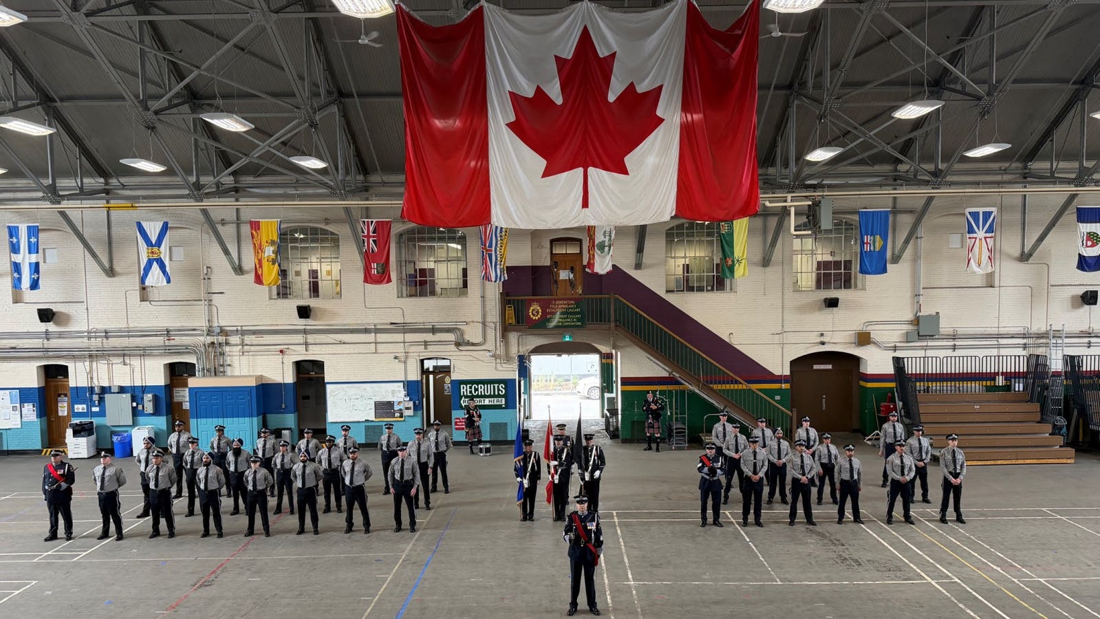 New transit peace officer recruits in gray uniforms stand at attention in formation inside a drill hall, with a color guard at center, a large Canadian flag hanging overhead and provincial and territorial flags lining the upper walls.
