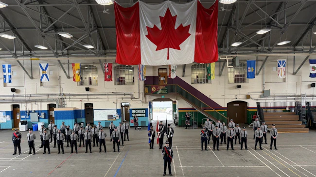 New transit peace officer recruits in gray uniforms stand at attention in formation inside a drill hall, with a color guard at center, a large Canadian flag hanging overhead and provincial and territorial flags lining the upper walls.