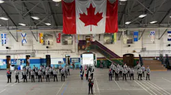 New transit peace officer recruits in gray uniforms stand at attention in formation inside a drill hall, with a color guard at center, a large Canadian flag hanging overhead and provincial and territorial flags lining the upper walls.