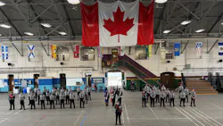 New transit peace officer recruits in gray uniforms stand at attention in formation inside a drill hall, with a color guard at center, a large Canadian flag hanging overhead and provincial and territorial flags lining the upper walls.