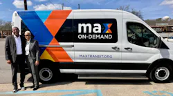 Two transit officials stand and smile beside a MAX On-Demand microtransit van bearing the maxtransit.org URL and a bold geometric wrap in blue, orange, and yellow. The Ford Transit-style vehicle is numbered MT2501 and parked in a lot with railroad tracks visible in the background.