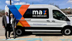 Two transit officials stand and smile beside a MAX On-Demand microtransit van bearing the maxtransit.org URL and a bold geometric wrap in blue, orange, and yellow. The Ford Transit-style vehicle is numbered MT2501 and parked in a lot with railroad tracks visible in the background.