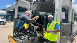 A transit driver in a yellow safety vest assists a wheelchair user boarding a microtransit van via a deployed rear wheelchair ramp, with the van's rear doors open wide and a transit station visible in the background.