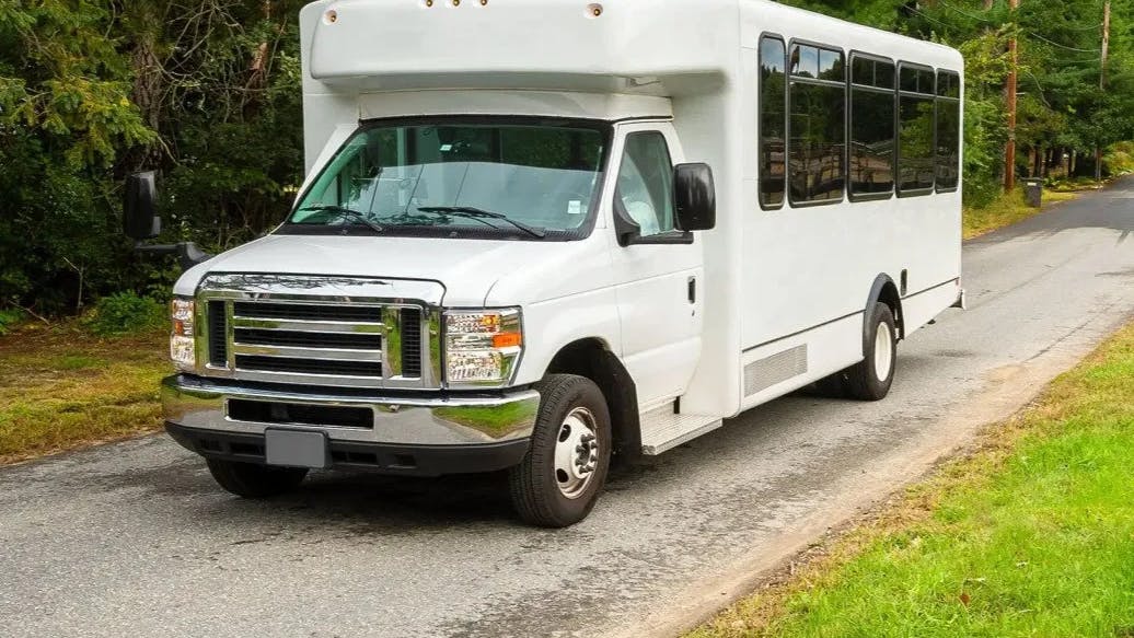A white Ford E-Series cutaway small transit bus traveling along a tree-lined rural road, with tinted passenger windows along the side and no agency markings visible.