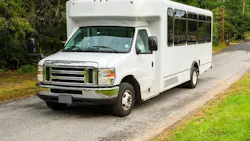 A white Ford E-Series cutaway small transit bus traveling along a tree-lined rural road, with tinted passenger windows along the side and no agency markings visible.