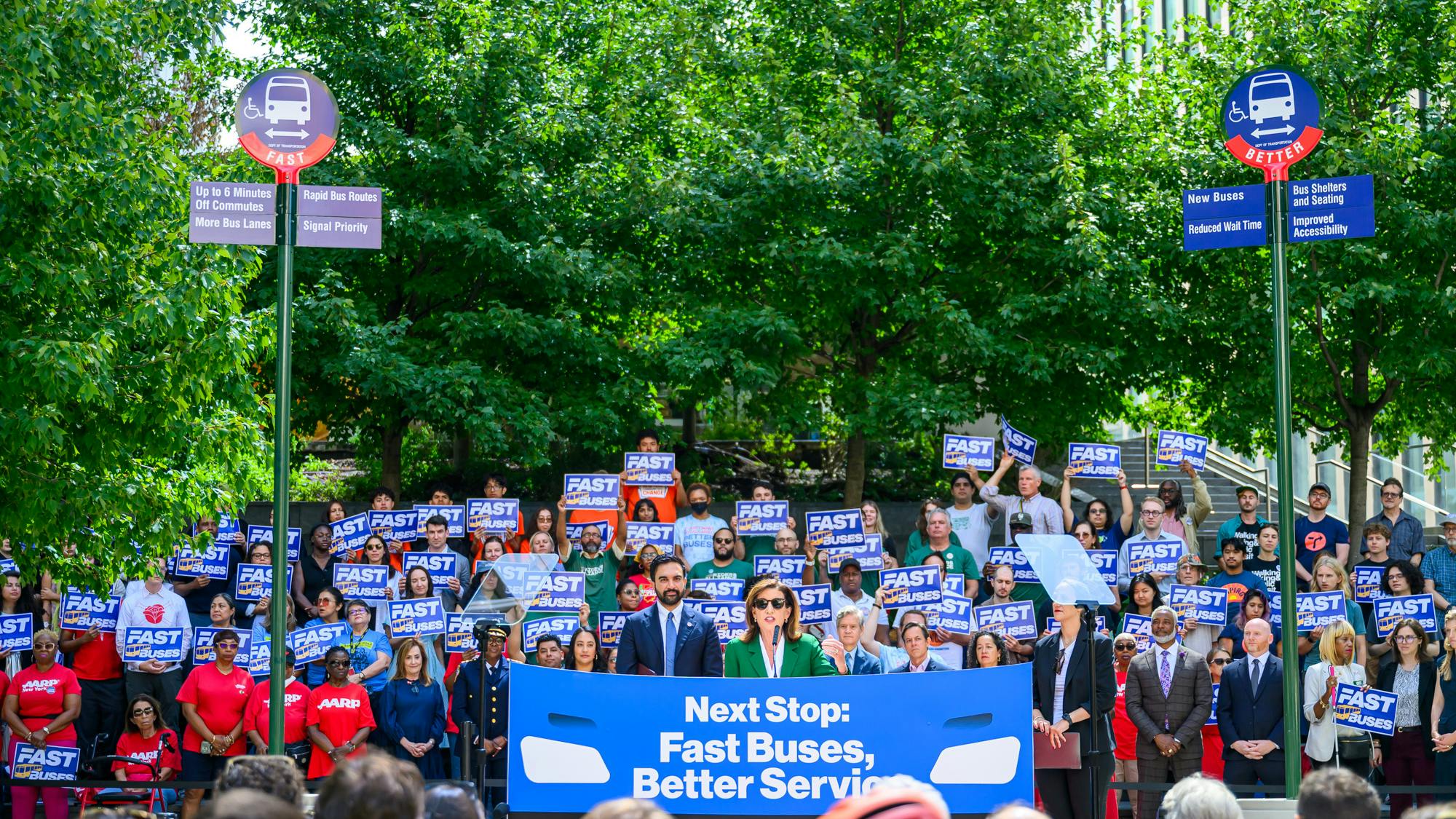New York City (NYC) Gov. Kathy Hochul and NYC Mayor Zohran Kwame Mamdani addresses a large outdoor rally in front of a crowd holding blue 'Fast Buses' signs, flanked by two mock bus stop poles listing bus rapid transit improvements such as signal priority, rapid bus routes, new buses, and reduced wait times.