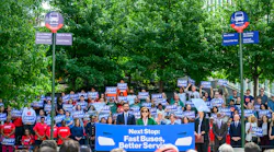 New York City (NYC) Gov. Kathy Hochul and NYC Mayor Zohran Kwame Mamdani addresses a large outdoor rally in front of a crowd holding blue 'Fast Buses' signs, flanked by two mock bus stop poles listing bus rapid transit improvements such as signal priority, rapid bus routes, new buses, and reduced wait times.