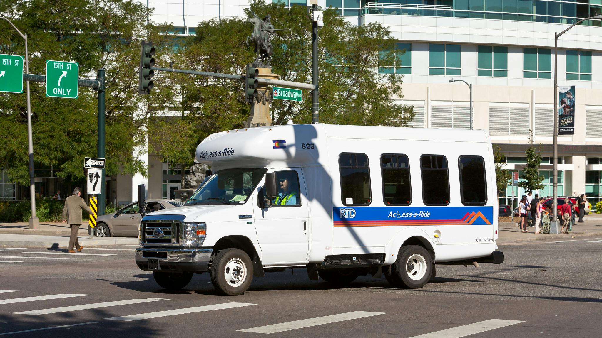 A Denver Regional Transportation District Access-a-Ride paratransit van number 623 traveling through the intersection of 15th Street and Broadway in downtown Denver, Colorado, displaying the agency's blue, red, and orange striped livery with the Colorado state flag emblem on a white Ford E-Series cutaway body.