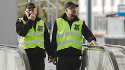 Two transit security officers in black uniforms and high-visibility yellow safety vests with badge insignia patrol an escalator at a transit station, one speaking into a radio while the other surveys the surroundings.