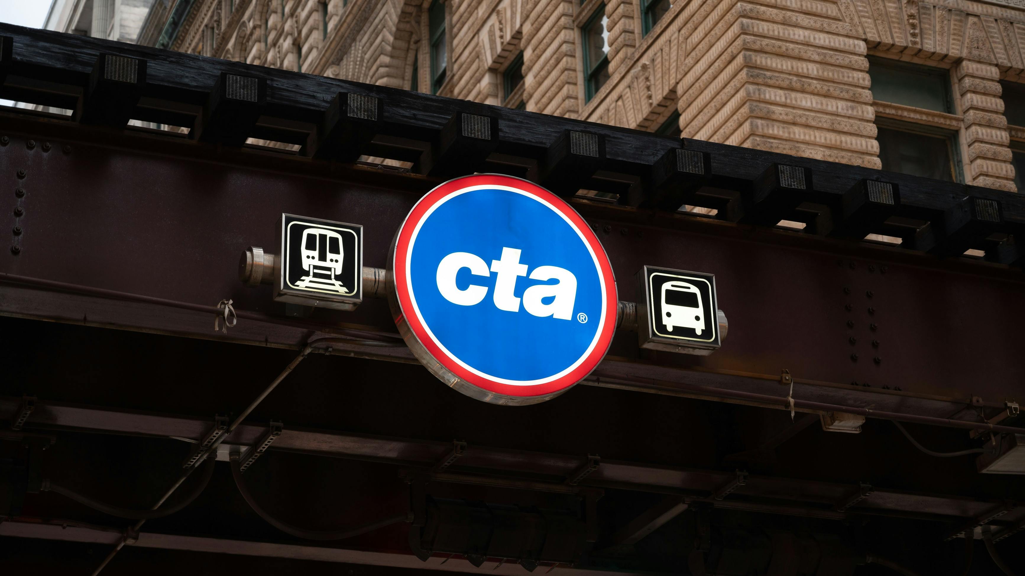 A circular blue and red Chicago Transit Authority sign with the 'cta' wordmark hangs beneath an elevated track structure, flanked by illuminated train and bus pictogram signs, with an ornate stone and brick building facade visible above.