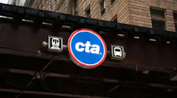 A circular blue and red Chicago Transit Authority sign with the 'cta' wordmark hangs beneath an elevated track structure, flanked by illuminated train and bus pictogram signs, with an ornate stone and brick building facade visible above.