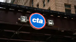A circular blue and red Chicago Transit Authority sign with the 'cta' wordmark hangs beneath an elevated track structure, flanked by illuminated train and bus pictogram signs, with an ornate stone and brick building facade visible above.