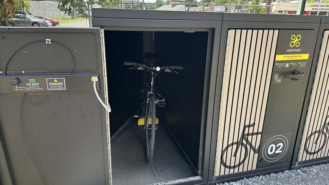 An open Velomodo secure bike locker at SEPTA's Fox Chase Regional Rail station reveals a bicycle stored inside, with an adjacent closed locker unit displaying the Velomodo logo, a bike icon and the number '02.'