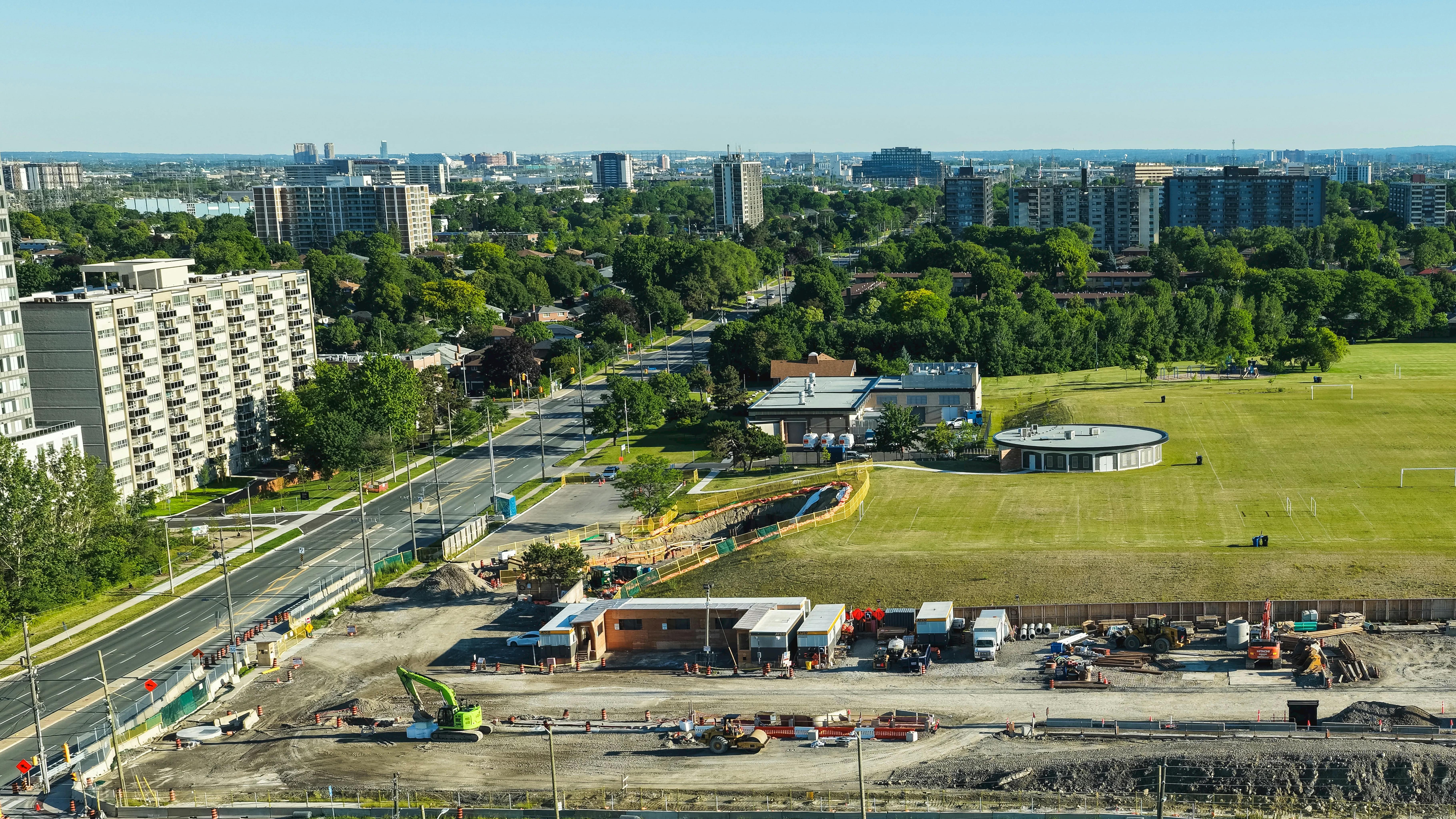 An aerial view of a Metrolinx Eglinton Crosstown West Extension construction site in Toronto, showing excavation work with piling rigs, cranes and support trailers alongside a busy multi-lane roadway, with a soccer field, apartment towers and the distant city skyline visible in the background.