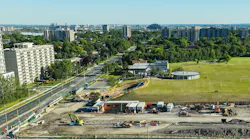 An aerial view of a Metrolinx Eglinton Crosstown West Extension construction site in Toronto, showing excavation work with piling rigs, cranes and support trailers alongside a busy multi-lane roadway, with a soccer field, apartment towers and the distant city skyline visible in the background.
