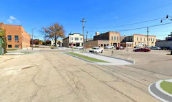 A street-level rendering overlaid on a Google Street View image of a Midwest commercial intersection, showing proposed curb extensions and green-painted pedestrian refuge islands added to an otherwise cracked and aging roadway, with brick storefronts and a two-story industrial building visible in the background.