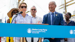 Massachusetts Gov. Maura Healey speaks at a podium alongside MBTA General Manager Phillip Eng and other officials during a ceremonial ribbon cutting for the MBTA's new Harbor Loop Ferry service, with a blue ceremonial ribbon in the foreground and the masts of a tall ship rising behind them.