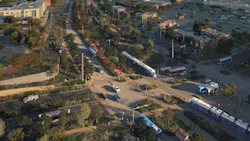 A rendering of Austin's future light rail line along Pleasant Valley Road, showing a modern white and blue light rail vehicle traveling alongside a landscaped multi-use path and roadway, with strip-mall retail and parking lots to the sides and the downtown Austin skyline in the distance at dusk.