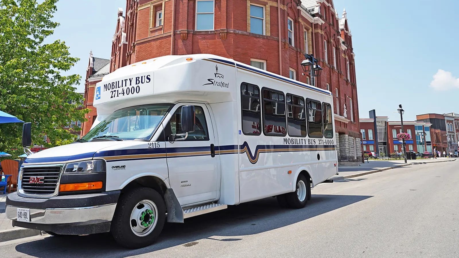 A white Stratford Mobility Bus paratransit vehicle parked on a downtown street in Stratford, Ontario, with a phone number displayed for booking and an ornate red-brick Victorian building with turrets and gables rising behind.