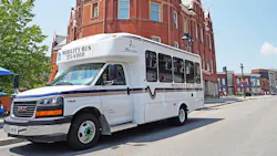 A white Stratford Mobility Bus paratransit vehicle parked on a downtown street in Stratford, Ontario, with a phone number displayed for booking and an ornate red-brick Victorian building with turrets and gables rising behind.
