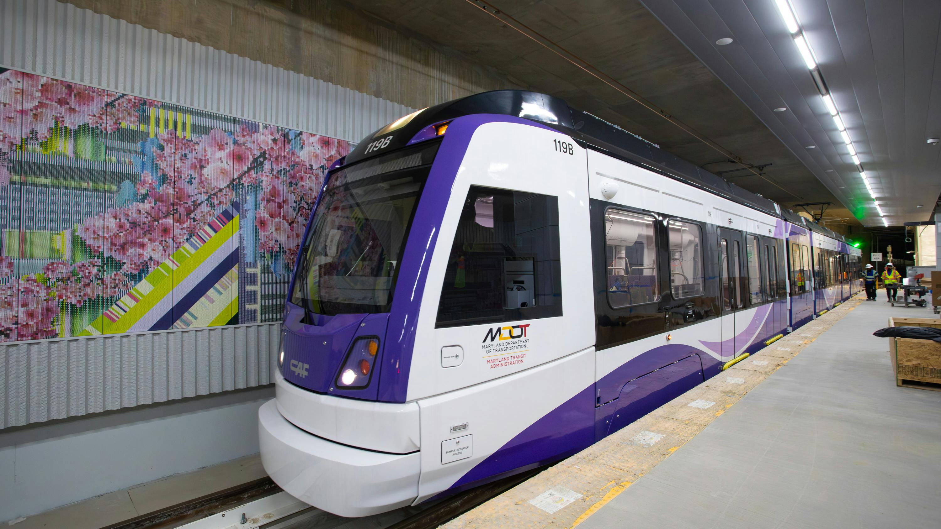 Maryland Transit Administration Purple Line light rail vehicle number 119B, a CAF-built car in white and purple livery, sits at the under-construction Bethesda Station platform, with a colorful cherry blossom-themed station mural visible on the tunnel wall to the left and construction workers visible in the background.