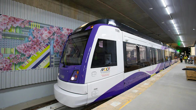 Maryland Transit Administration Purple Line light rail vehicle number 119B, a CAF-built car in white and purple livery, sits at the under-construction Bethesda Station platform, with a colorful cherry blossom-themed station mural visible on the tunnel wall to the left and construction workers visible in the background.
