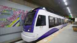 Maryland Transit Administration Purple Line light rail vehicle number 119B, a CAF-built car in white and purple livery, sits at the under-construction Bethesda Station platform, with a colorful cherry blossom-themed station mural visible on the tunnel wall to the left and construction workers visible in the background.