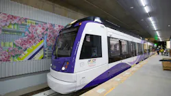 Maryland Transit Administration Purple Line light rail vehicle number 119B, a CAF-built car in white and purple livery, sits at the under-construction Bethesda Station platform, with a colorful cherry blossom-themed station mural visible on the tunnel wall to the left and construction workers visible in the background.