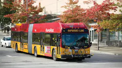 King County Metro RapidRide I Line articulated hybrid-electric bus number 6020 in the signature red and yellow RapidRide livery, displaying 'RapidRide I Line' on its destination board and carrying a bicycle on its front rack, traveling along an urban street lined with red-leafed autumn trees.