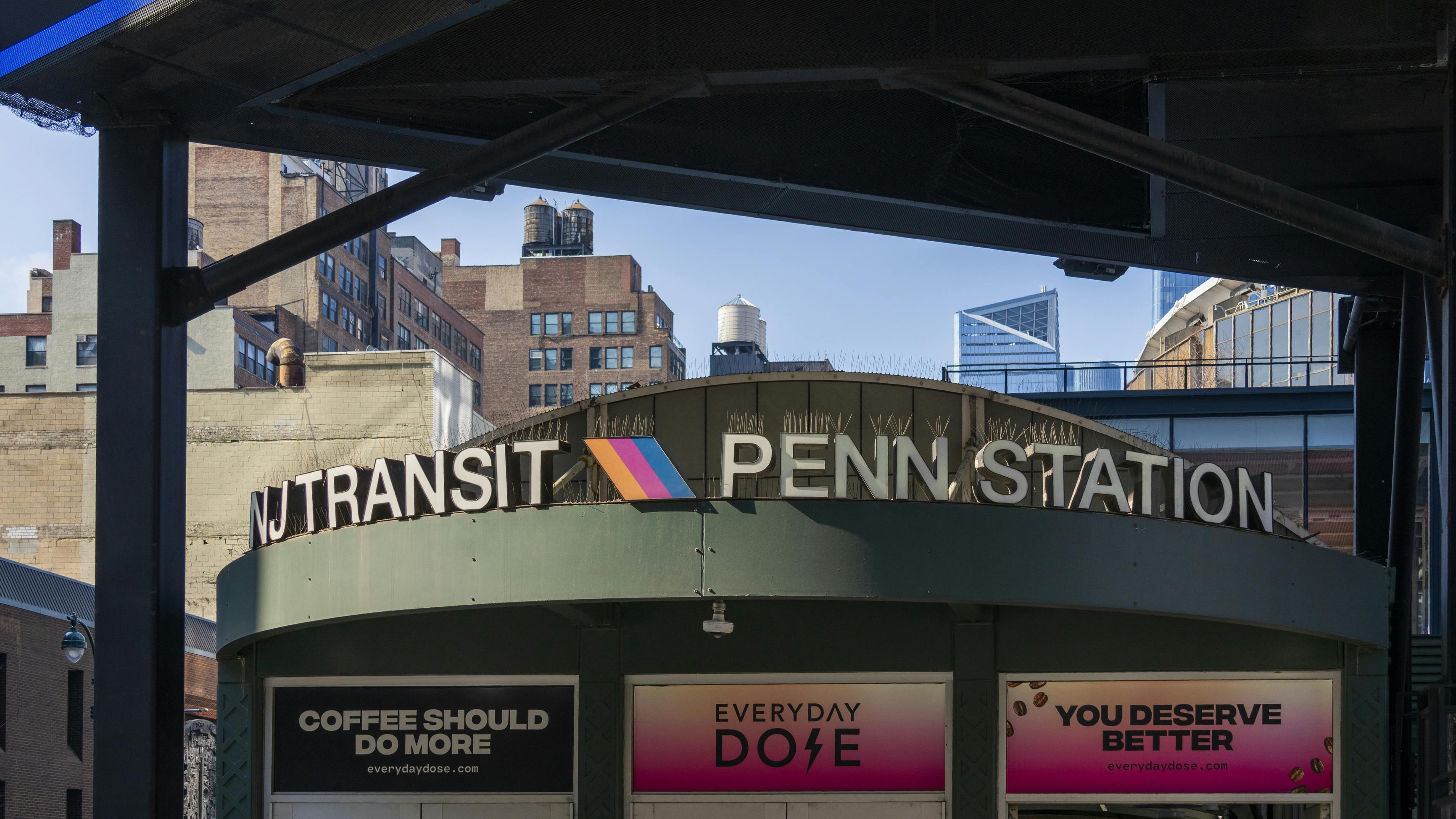 A rounded green entrance kiosk marked 'NJ Transit Penn Station' with the agency's colorful chevron logo sits beneath a modern black steel canopy in Midtown Manhattan, with brick buildings, rooftop water towers and the Hudson Yards skyline visible in the background.