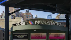A rounded green entrance kiosk marked 'NJ Transit Penn Station' with the agency's colorful chevron logo sits beneath a modern black steel canopy in Midtown Manhattan, with brick buildings, rooftop water towers and the Hudson Yards skyline visible in the background.