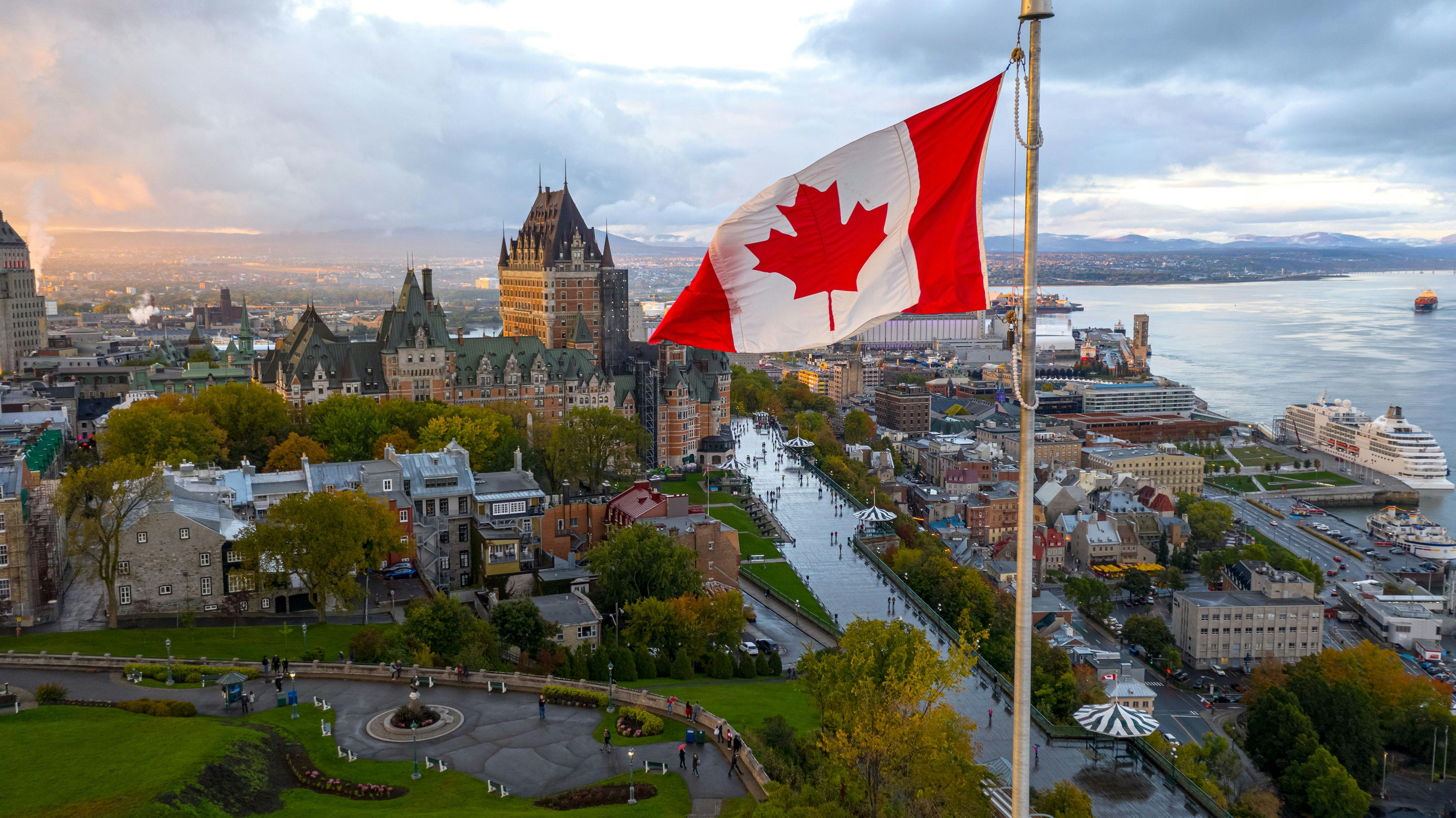 An aerial view of Old Quebec City at dusk, with the Canadian flag flying in the foreground, the Ch&acirc;teau Frontenac castle hotel dominating the historic skyline, the St. Lawrence River and its port visible in the background, and autumn foliage surrounding the UNESCO World Heritage site.