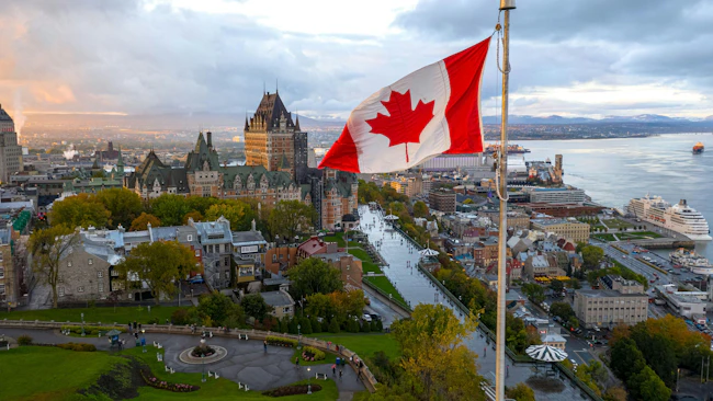 An aerial view of Old Quebec City at dusk, with the Canadian flag flying in the foreground, the Ch&acirc;teau Frontenac castle hotel dominating the historic skyline, the St. Lawrence River and its port visible in the background, and autumn foliage surrounding the UNESCO World Heritage site.