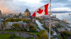 An aerial view of Old Quebec City at dusk, with the Canadian flag flying in the foreground, the Château Frontenac castle hotel dominating the historic skyline, the St. Lawrence River and its port visible in the background, and autumn foliage surrounding the UNESCO World Heritage site.