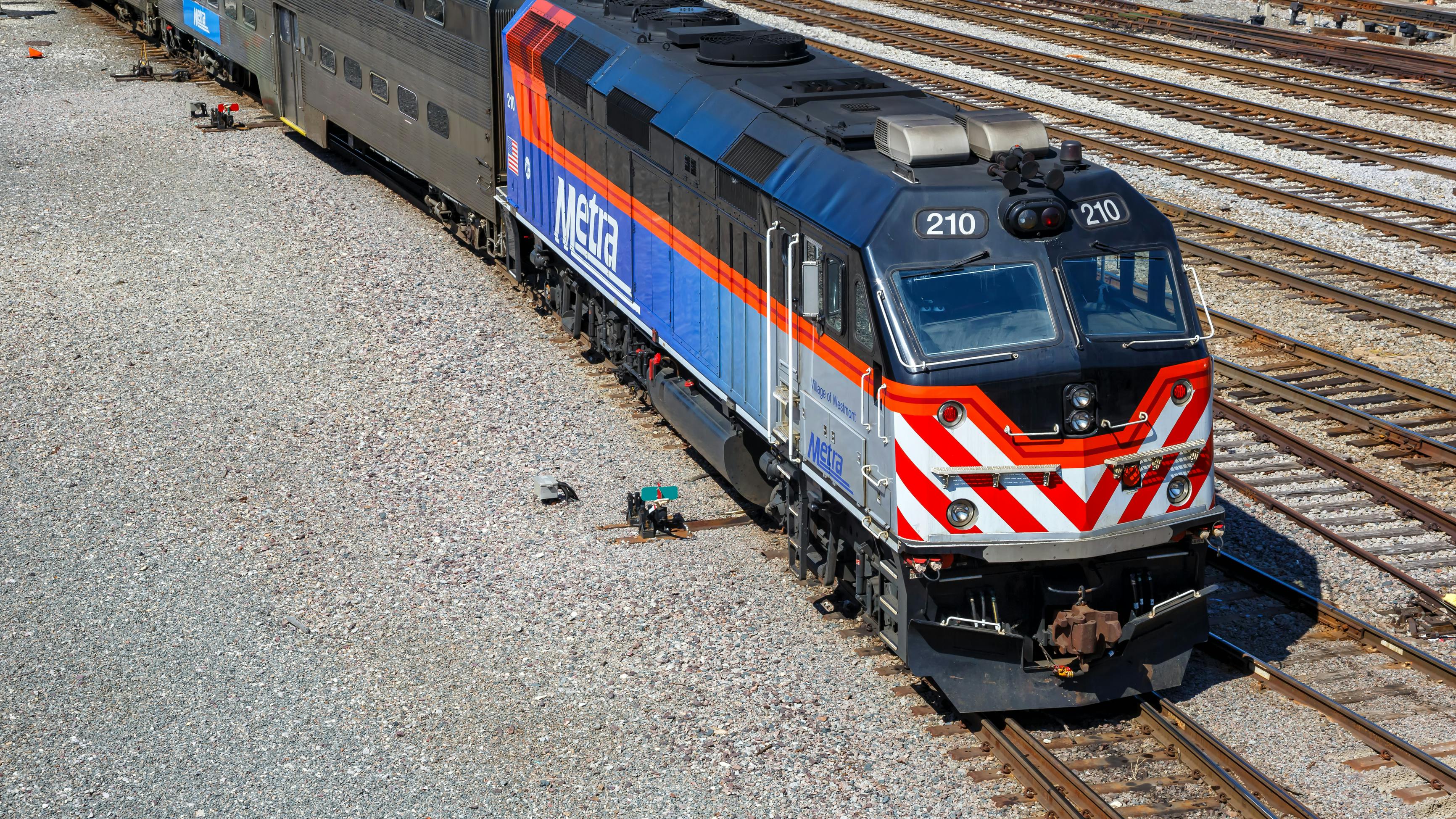 An overhead view of a blue and orange Metra commuter rail F40 locomotive numbered 210 sitting on track in a Chicago rail yard, coupled to bilevel gallery coaches, with additional tracks and equipment visible around it.