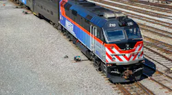 An overhead view of a blue and orange Metra commuter rail F40 locomotive numbered 210 sitting on track in a Chicago rail yard, coupled to bilevel gallery coaches, with additional tracks and equipment visible around it.