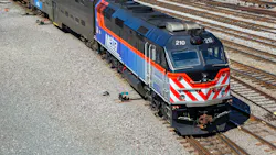 An overhead view of a blue and orange Metra commuter rail F40 locomotive numbered 210 sitting on track in a Chicago rail yard, coupled to bilevel gallery coaches, with additional tracks and equipment visible around it.