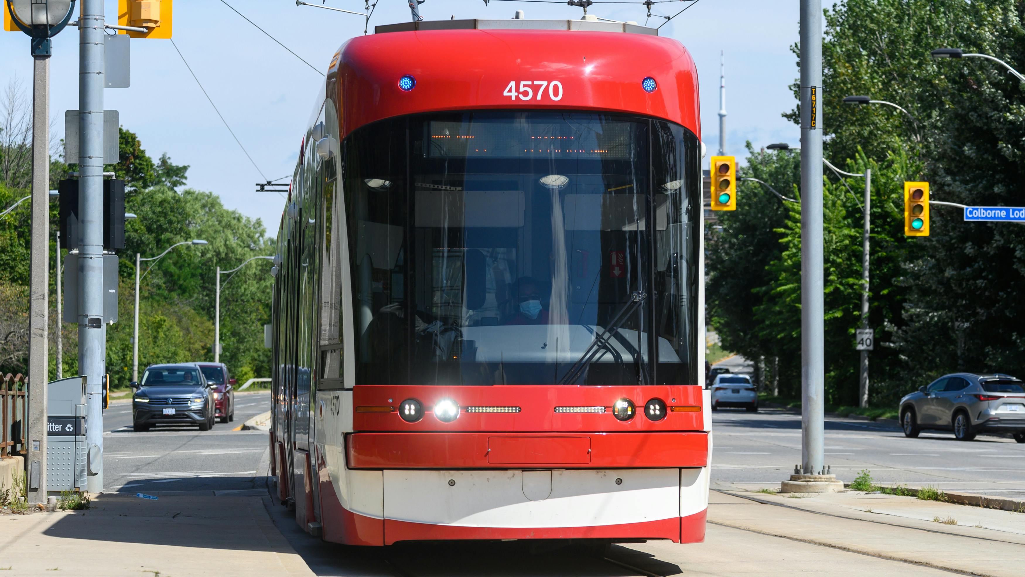 A red and white Toronto Transit Commission Flexity streetcar numbered 4570 travels along a street-level dedicated right-of-way in Toronto, with the CN Tower visible in the distance and traffic signals and trees flanking the tracks.
